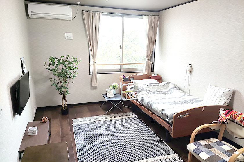 窓採光が良い介護施設の居室。ベッド、テレビ、観葉植物が配置されている。